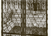 This postcard depicts a close up of two panels of a grille or iron gate that is located in the House of Commons, London. Through the cast iron you can see an empty house of parliament with wooden benches and ornate decoration.