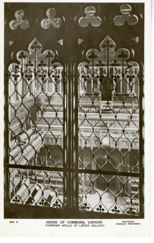 This postcard depicts a close up of two panels of a grille or iron gate that is located in the House of Commons, London. Through the cast iron you can see an empty house of parliament with wooden benches and ornate decoration.