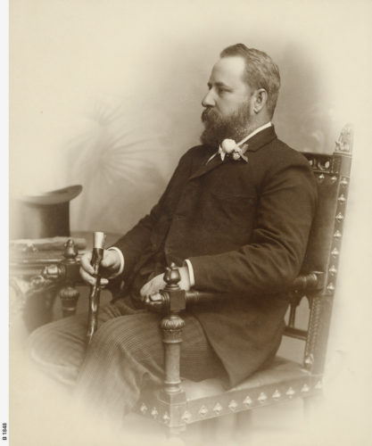 A portrait of Charles Cameron Kingston. He is sitting in a decoratively carved arm chair looking to his left. He has a carved wooden cane in his right hand. There is a top hat sitting on a small table to the right hand side of the chair. He has a full head of hair and a full mustache and beard.