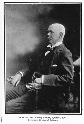 A portrait of Josiah Symon sitting in an arm chair facing his left. He is wearing a formal suit, and the background in neutral. He is a blad man, but with a thick mustache. 