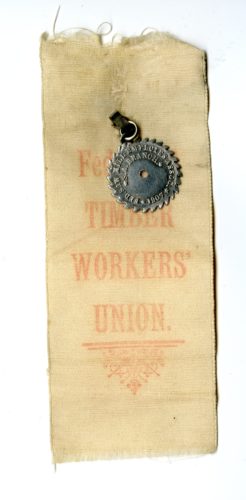 An off white, almost yellow, fabric ribbon in a long vertical rectangle with a small metal disk with spiked edged pinned to the top centre. The ribbon read, in red text, Federated Timber Workers Union.