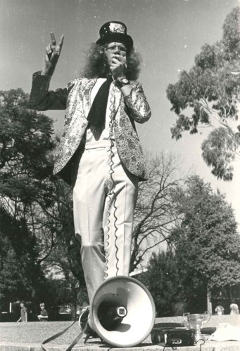 A young man with clurly hair in a top had with glittery blazer and while slacks standing in a park with a megaphone giving a speech
