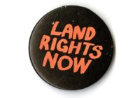 A black badge with red text that says LAND RIGHTS NOW.