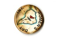 A small discolored white badge whihc the words INT. Women's Day written on it in black text. In the Centre if an outline of South America with a rose inside of it.