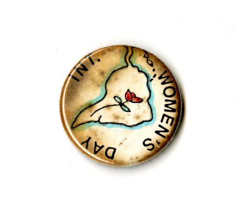 A small discolored white badge whihc the words INT. Women's Day written on it in black text. In the Centre if an outline of South America with a rose inside of it.
